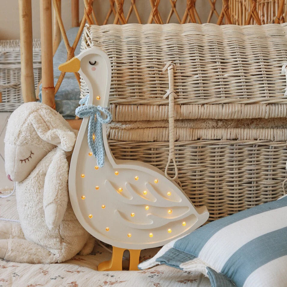 A cozy scene with a wicker chest in the background. In front of it is a plush toy lamb lying down, and next to it is a handmade wooden goose lamp adorned with small glowing bulbs and a blue ribbon around its neck. The nursery decor setting is soft and inviting.