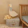 A Handmade Wooden Goose Lamp, complete with small circular cutouts and a light blue bow around its neck, stands on a white wicker basket. A bunch of beige fabric flowers lies beside the nursery decor. The charming scene is set beside a wicker chair against a beige wall.