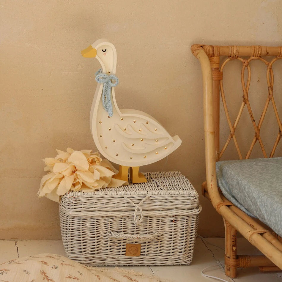 A Handmade Wooden Goose Lamp, complete with small circular cutouts and a light blue bow around its neck, stands on a white wicker basket. A bunch of beige fabric flowers lies beside the nursery decor. The charming scene is set beside a wicker chair against a beige wall.