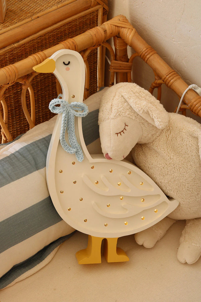 A cozy scene with a stuffed lamb and a nursery decor marvel—a Handmade Wooden Goose Lamp. The white, plush lamb lies against a striped fabric pillow, while the cream-colored goose light, adorned with small bulbs and a blue knitted bow, rests on a woven wooden chair. Perfect children's night light ambiance.