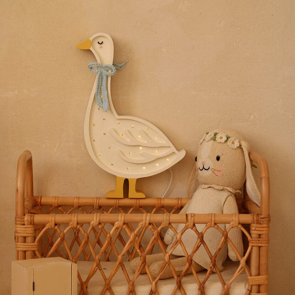 A cozy scene featuring a wicker bassinet with a plush bunny toy, wearing a floral headband, resting inside. Above, a Handmade Wooden Goose Lamp adds warmth to the light beige backdrop, enhancing the nursery decor perfectly.