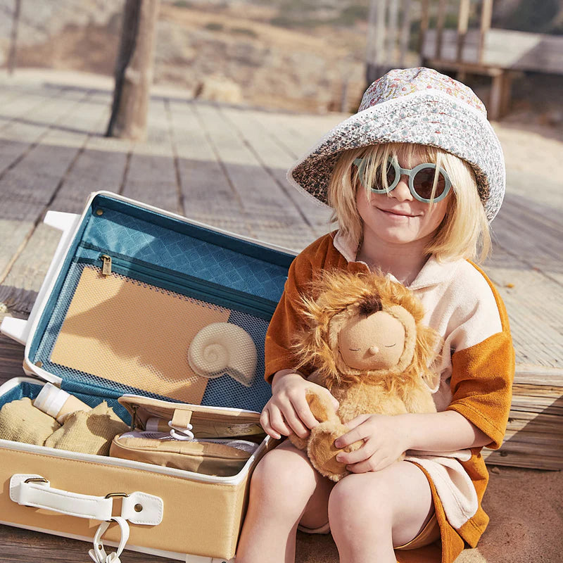 A young child in sunglasses and a wide-brimmed hat sits on a wooden boardwalk, holding a stuffed lion toy, with an open Olli Ella See-Ya Suitcase - Butterscotch beside them.