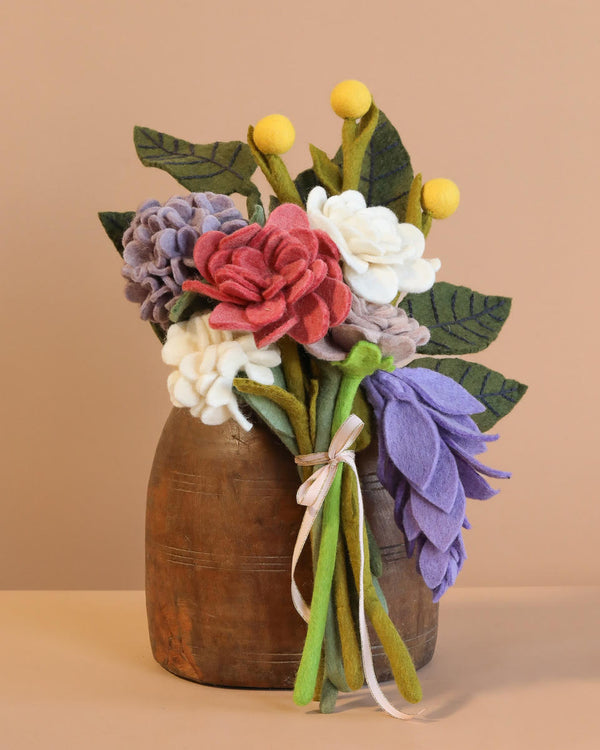 The Handcrafted Felt Flower Bouquet features wool-felt blooms in purple, pink, white, and green nestled in a rustic wooden vase. Yellow accents complement the arrangement against a warm beige backdrop, enhancing its cozy, handmade charm.
