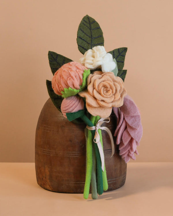 The Handcrafted Felt Flower Bouquet showcases pink, peach, and white blooms elegantly arranged in a wooden vase against a beige background. Green felt stems and leaves are tied with a ribbon, creating a sustainable floral decor piece.