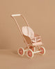 The Maileg Stroller, Micro - Powder is a small, pastel pink stroller with white wheels and a polka-dot fabric seat, shown against a beige background.