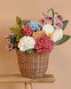 The Basket Of Felt Flowers features a wicker basket filled with a handmade floral arrangement of vibrant felt flowers in pink, red, white, and blue hues with green leaves. This sustainable display includes a charming felt hydrangea set against a beige background.