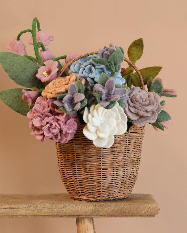 The Basket Of Felt Flowers features a handmade floral arrangement with colorful felt roses, sweet pea blooms, and hydrangeas in soft pink, cream, lavender, and blue shades. This sustainable bouquet is displayed in a wicker basket on a wooden surface against a beige background.