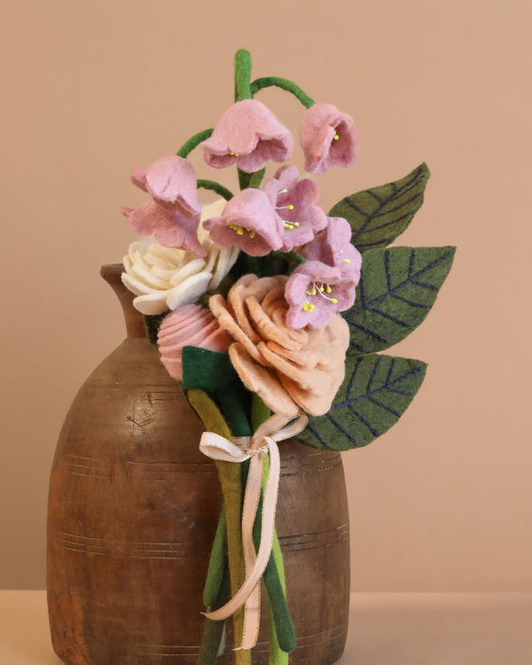 The Handcrafted Felt Flower Bouquet features pink and cream blooms with green leaves in a textured brown vase. These eco-friendly florals have delicate petals, elegantly tied with a ribbon, set against a neutral background.