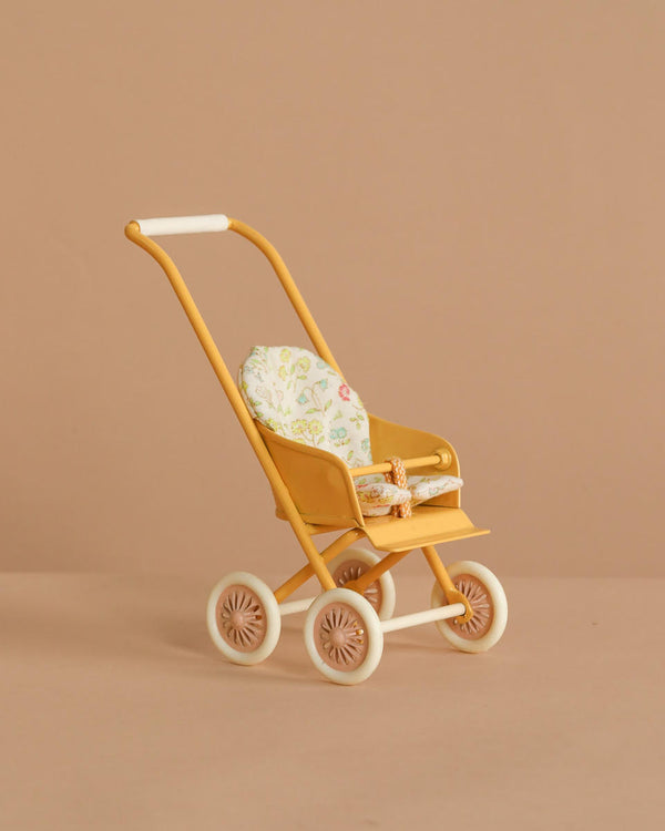 The Maileg Stroller, Micro - Yellow features a vintage design with white wheels, a floral seat cushion, and an orange-and-white safety strap, shown against a plain beige background.