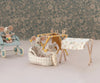 A whimsical scene featuring baby mice: one sits in a baby carriage holding a bottle, another is in a small bassinet with adorable baby accessories, while a third hangs clothes on a tiny clothesline by the **Maileg Nursery Table, Baby Mouse - Ocher**. The backdrop is a soft floral pattern, creating a cozy atmosphere.