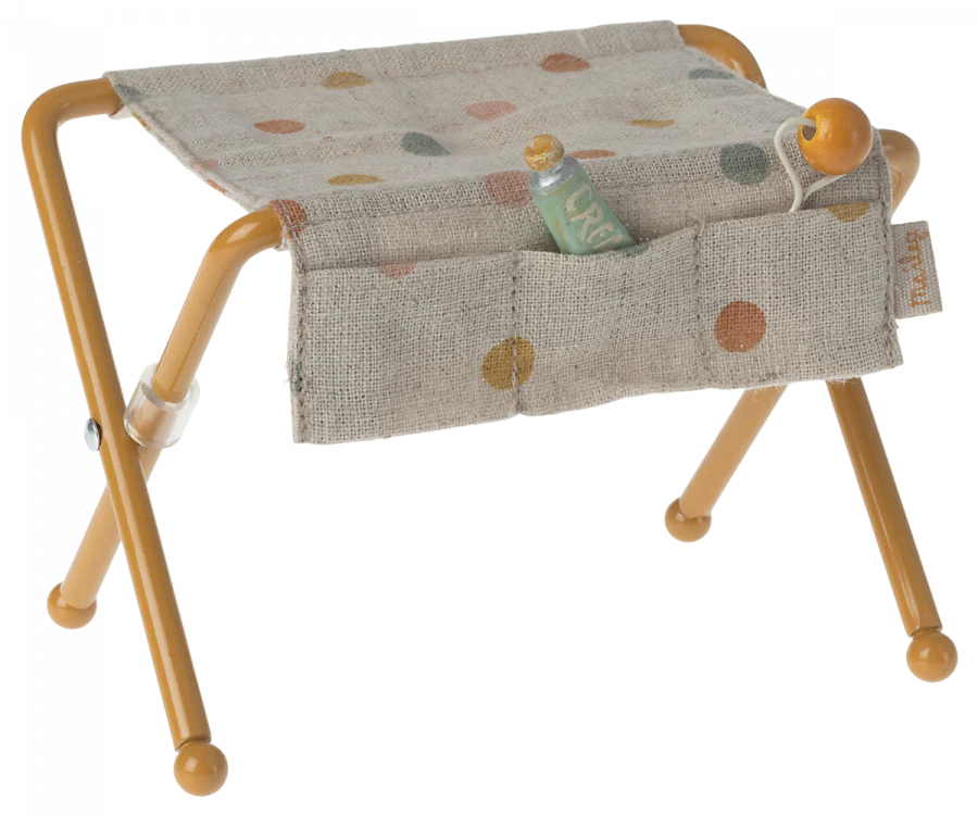 A small, foldable stool with yellow metal legs and a fabric seat featuring multicolored dots. Perfect for a **Maileg Nursery Table, Baby Mouse - Ocher**, it comes with a fabric pouch holding baby accessories like a tiny green bottle and a small orange ball.