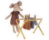 A plush mouse from the mouse family is dressed in a knitted dress, standing beside a small yellow Maileg Drying Rack, Mouse Size. The rack holds two pieces of doll-sized clothing secured with small wooden pegs.