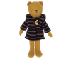 Teddy Junior, dressed in a light brown hue, wears the Maileg Extra Clothing: Duffle Coat featuring a dark blue hood with beige horizontal stripes and a toggle button. The plush teddy bear stands upright and has simple facial features with stitched eyes and nose.