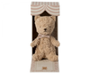 A Maileg My First Teddy, Powder is seated inside an open, striped cardboard box with an arch-like design at the top. The cream-colored bear, adorned with a bow, features black eyes and nose. This vintage teddy bear makes a perfect personalized baby keepsake or new born baby gift, complete with decorative elements.