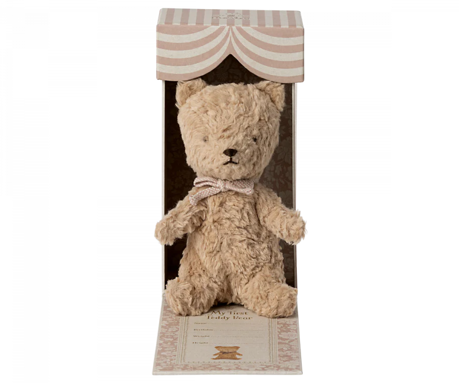 A Maileg My First Teddy, Powder is seated inside an open, striped cardboard box with an arch-like design at the top. The cream-colored bear, adorned with a bow, features black eyes and nose. This vintage teddy bear makes a perfect personalized baby keepsake or new born baby gift, complete with decorative elements.