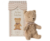 A plush teddy bear with light brown fur and a ribbon around its neck stands next to a decorative, vintage-style box that reads "Maileg My First Teddy, Powder" with an illustration of the same bear. The box, perfect for a new born baby, has a striped, circus-tent-like design at the top.