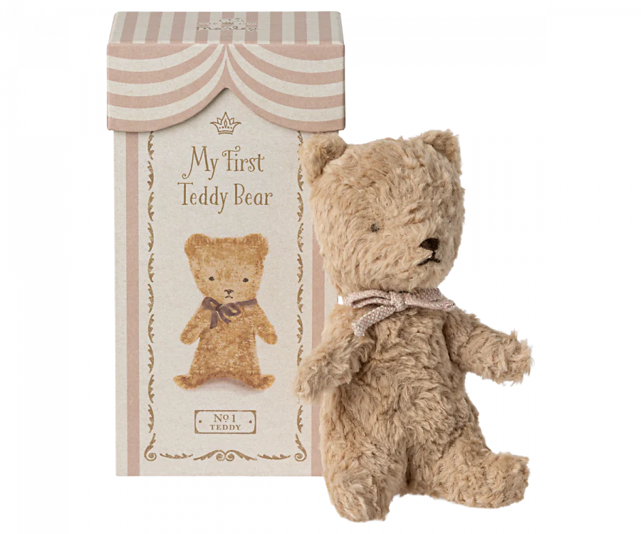 A plush teddy bear with light brown fur and a ribbon around its neck stands next to a decorative, vintage-style box that reads "Maileg My First Teddy, Powder" with an illustration of the same bear. The box, perfect for a new born baby, has a striped, circus-tent-like design at the top.