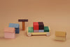 Raduga Grez Vehicle Blocks, crafted from solid wood and painted with eco-friendly colors, are arranged on a brown background to resemble a racing car, two small houses, a tall structure, and a cube in vibrant shades of yellow, red, green, blue, and orange for safe play.