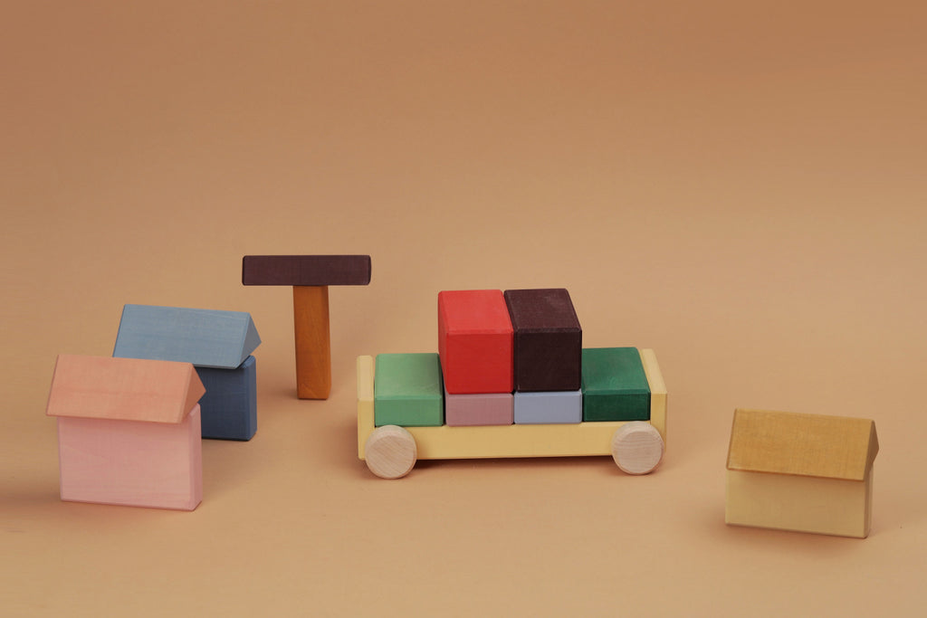 Raduga Grez Vehicle Blocks, crafted from solid wood and painted with eco-friendly colors, are arranged on a brown background to resemble a racing car, two small houses, a tall structure, and a cube in vibrant shades of yellow, red, green, blue, and orange for safe play.