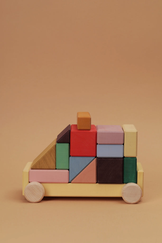 The Raduga Grez Vehicle Blocks form a geometric toy racing car crafted from solid wood, arranged on a light brown background. The eco-friendly painted blocks are in shades of green, red, mustard, blue, and purple with round wheels.