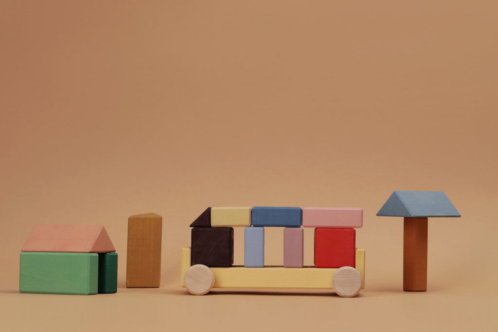Raduga Grez Vehicle Blocks feature pastel wooden pieces arranged on a beige background, forming a house, tall tower, racing car with wheels, and tree with brown trunk and blue top. Made from solid wood and eco-friendly paint, these blocks spark creativity.
