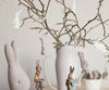 A cozy scene featuring a white ceramic pitcher holding bare branches adorned with Maileg Easter Bunny Ornaments, 5 pcs. Plush and ceramic rabbit figurines in various poses are arranged around the pitcher on a light wooden surface, creating a whimsical Easter ambiance.