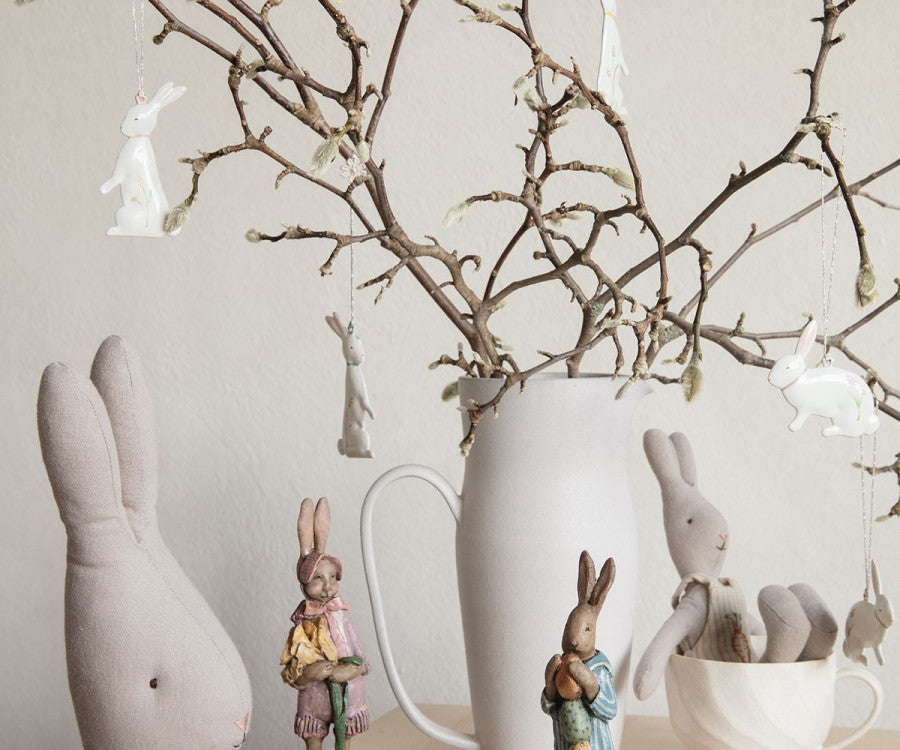 A cozy scene featuring a white ceramic pitcher holding bare branches adorned with Maileg Easter Bunny Ornaments, 5 pcs. Plush and ceramic rabbit figurines in various poses are arranged around the pitcher on a light wooden surface, creating a whimsical Easter ambiance.