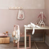 A children's room with soft pink and beige tones. A table with a white plush bunny, aptly named Maileg Bunny Bob, sits beside a pale pink chair. A coat hook holds a baby dress and a polka dot bag. A stuffed bear with a pink sweater is on a stand, and a doll bed is nearby.