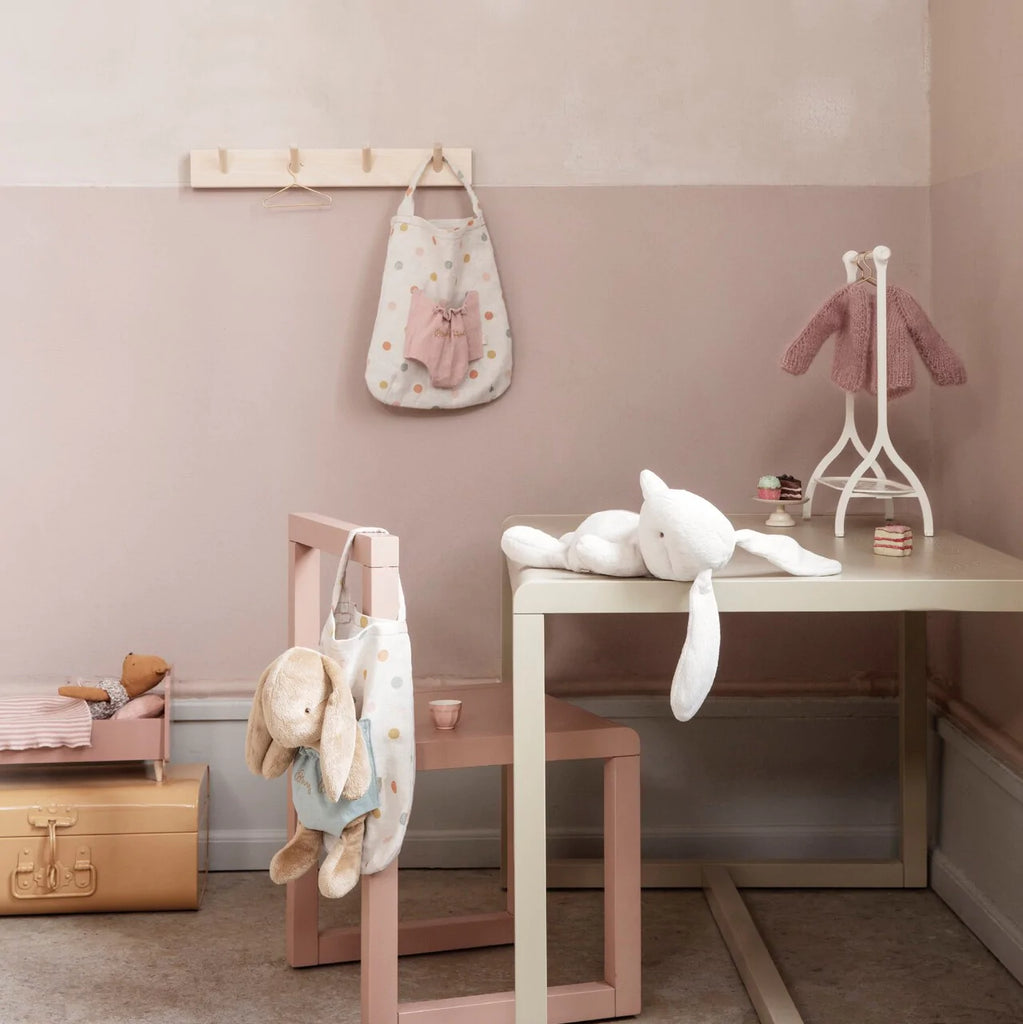 A children's room with soft pink and beige tones. A table with a white plush bunny, aptly named Maileg Bunny Bob, sits beside a pale pink chair. A coat hook holds a baby dress and a polka dot bag. A stuffed bear with a pink sweater is on a stand, and a doll bed is nearby.