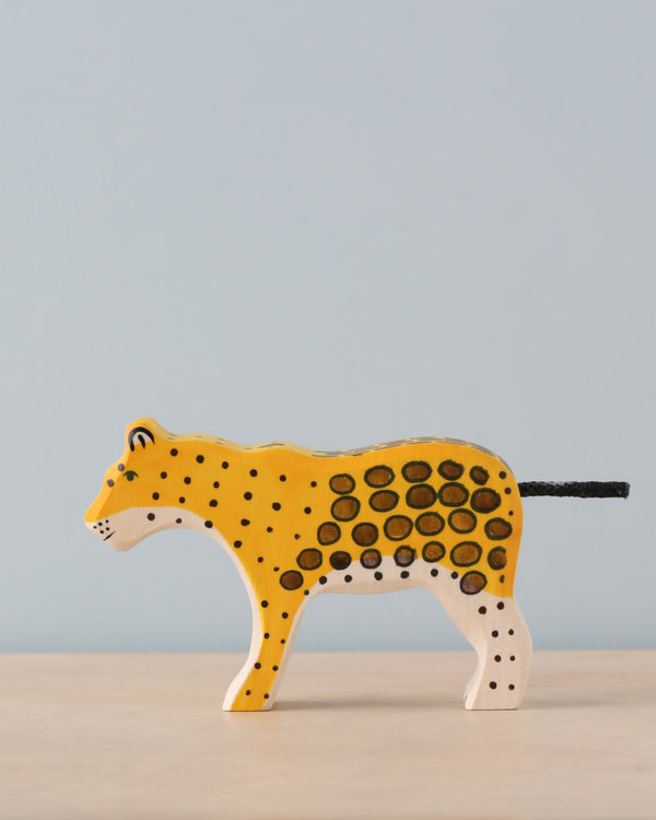 The Holztiger Leopard, a finely handcrafted wood toy from the exclusive HOLZTIGER collection, features a yellow body with black spots, elegantly posed on a light surface against a plain, light blue background—representing exceptional European craftsmanship.