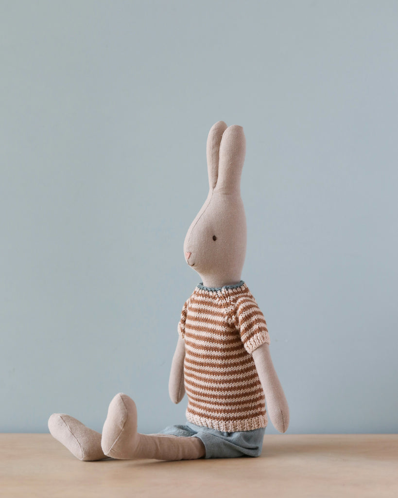 The Maileg Rabbit Size 3, Classic - Knitted Shirt and Shorts sits upright on a light-colored surface against a pale blue background. Made from soft natural linen, the rabbit wears a striped brown and white sweater paired with light blue shorts, with its legs extended in front. Its ears are upright, showcasing a simple, minimalist design.