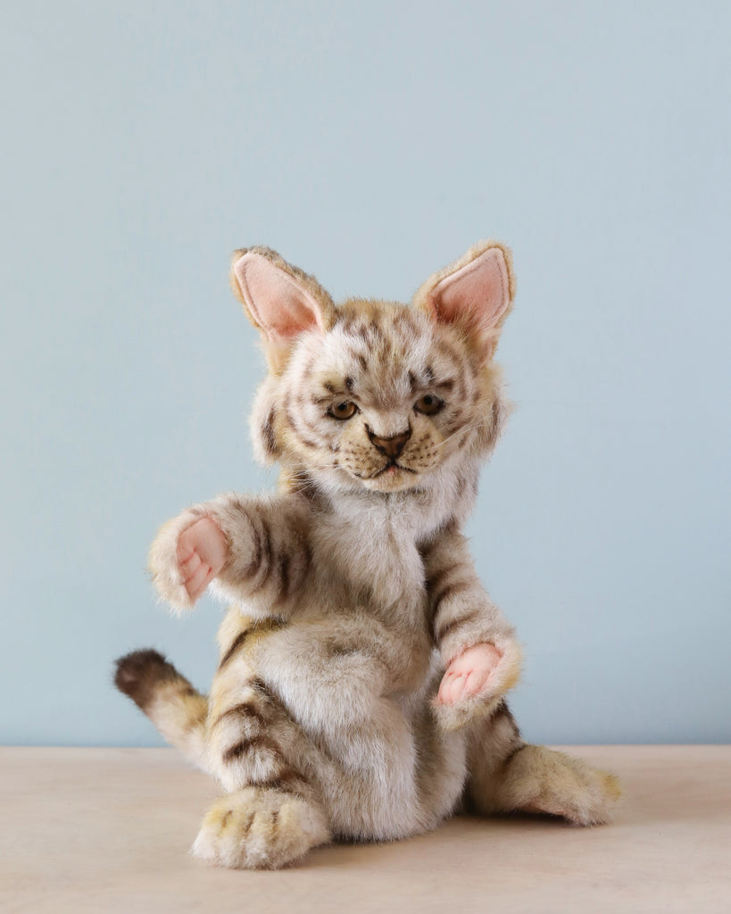 A Leopard Cat Puppet with realistic features sits upright on a wooden surface against a light blue background, showcasing its unique plush design and striped fur with both front paws raised.