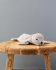 A Senger Naturwelt Cuddly Animal - Gray Seal lying on a rustic wooden stool against a plain grey backdrop. The seal is light grey with a simple, cute face.
