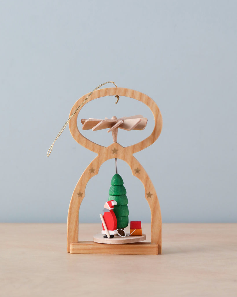 The Richard Glaesser Mini Pyramid Hanging Ornament - Santa, made in Germany, features a green tree with Santa on a sled, a propeller, and star details. It rests on light wood against a pale blue background.