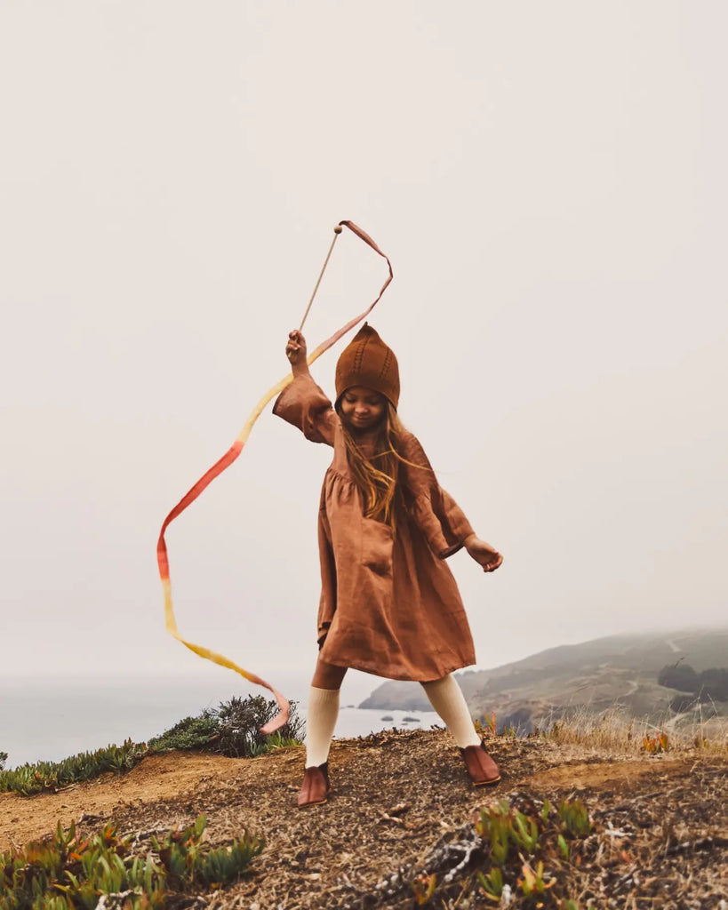 A young child wearing a brown hooded dress and white tights joyfully waves a Sarah's Silk Boho Streamer Wand on a foggy hillside. The background showcases a misty, coastal landscape with steep cliffs and sparse vegetation. The child appears carefree and engaged in open-ended play.