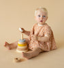 A toddler with blonde hair and blue eyes sits on a beige background, wearing a dotted dress and playing with a Raduga Grez Handmade Pyramid Tower Stacker in Sand.