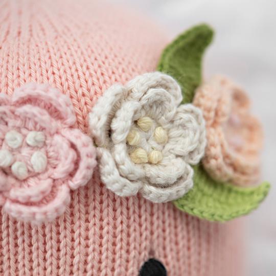 Close-up of a Cuddle + Kind Penelope the Flamingo adorned with crocheted flowers in white and pale peach, with green leaves and small yellow centers. Detailed and textured.