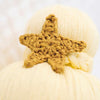 A close-up image of a hand-knit, golden crocheted Cuddle + Kind Skye the Mermaid attached to a creamy-white textured surface, possibly yarn or soft fabric, with a focus on the intricate details of the star's.