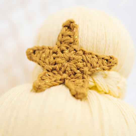 A close-up image of a hand-knit, golden crocheted Cuddle + Kind Skye the Mermaid attached to a creamy-white textured surface, possibly yarn or soft fabric, with a focus on the intricate details of the star's.