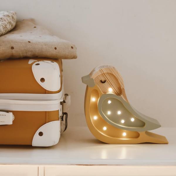 A cozy scene features a handmade pinewood lamp with small bulbs, emitting a warm glow. The Little Lights Bird Lamp is placed on a light-colored surface next to a mustard-colored suitcase adorned with white animal illustrations, with folded beige fabric on top.