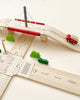 A Wooden Road System Deluxe set depicting a roadways scene with a bridge, a red toy car, three green bushes, and traffic signs on a light beige background.