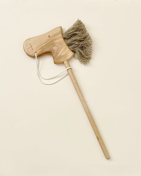 The Handmade Wooden Riding Horse, crafted from alder wood, features a carved head with simple facial features and a thick beige fiber mane. It includes a long wooden handle and a white string leash on a plain light background.