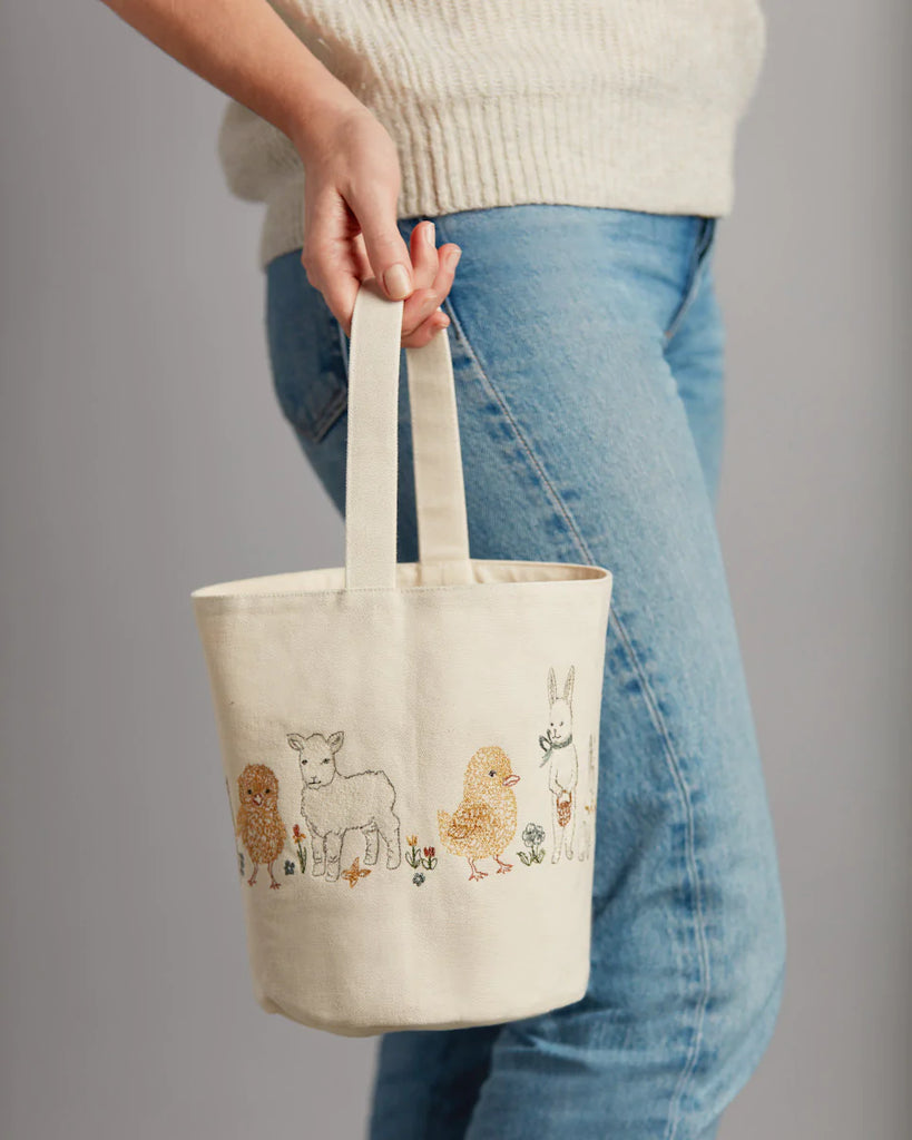 A person holds a Coral & Tusk Spring Babies Canvas Bucket embroidered with farm animals like a lamb, chick, and rabbit. Resembling an Easter basket, this charming tote pairs perfectly with their blue jeans and beige top against a plain gray background.