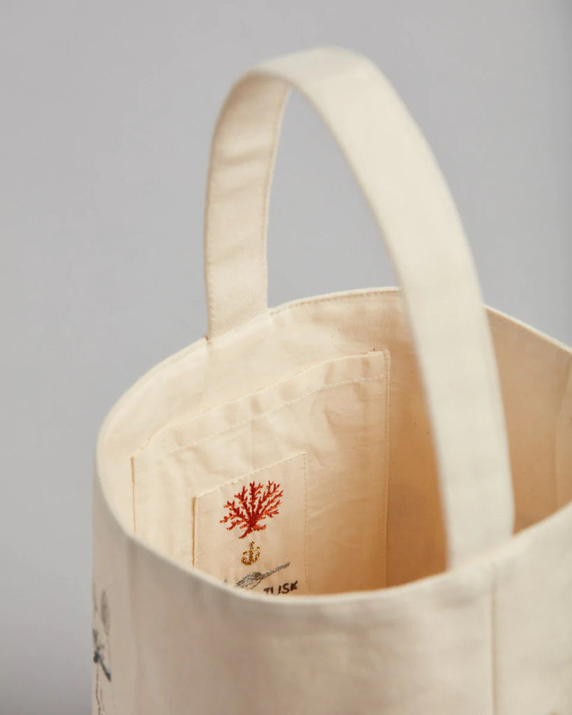 The Coral & Tusk Spring Babies Canvas Bucket features a beige canvas with an embroidered strap. Inside, delicate red coral designs add flair. Its textured finish and neutral gray background highlight its handmade charm, making it perfect as a unique Easter basket alternative.