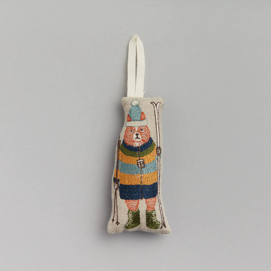 The Coral & Tusk Slopes Fox Ornament is a fabric decoration shaped like a bear wearing a colorful striped parka, green boots, and a cheerful pom-pom cap while holding ski poles. It features a loop for hanging against light gray.