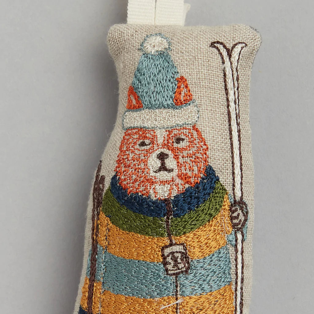 Embroidered ornament featuring a bear in a teal pom-pom cap, holding skis, wearing an orange, blue, and yellow striped jacket on light gray. Product: Coral & Tusk Slopes Fox Ornament.