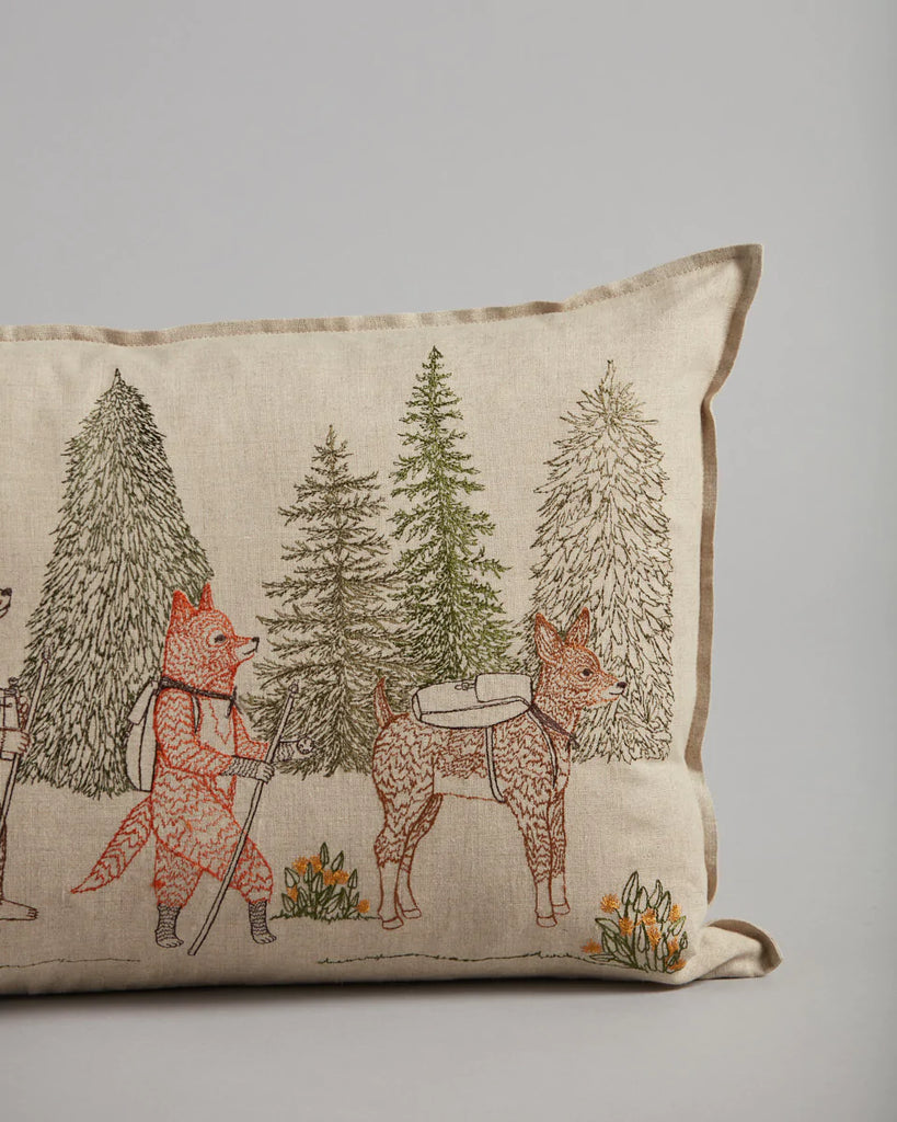 The Coral & Tusk Hikers Pillow is a beige cushion with embroidered foxes in hiking gear, trekking poles, evergreen trees, bags, and surrounded by orange flowers. The light gray background adds to its Camp Coral & Tusk charm. Ships in approximately one week.