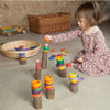 A young child plays with Grapat 36 rings in 6 colors stacked on pegs, sitting on a concrete floor surrounded by a wicker basket and a wooden toy truck.