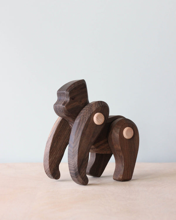 The Handmade Wooden Gorilla toy boasts movable limbs, crafted expertly from dark and light wood. It stands on a wooden surface against a light blue backdrop, capturing the charm of artisanal toys.