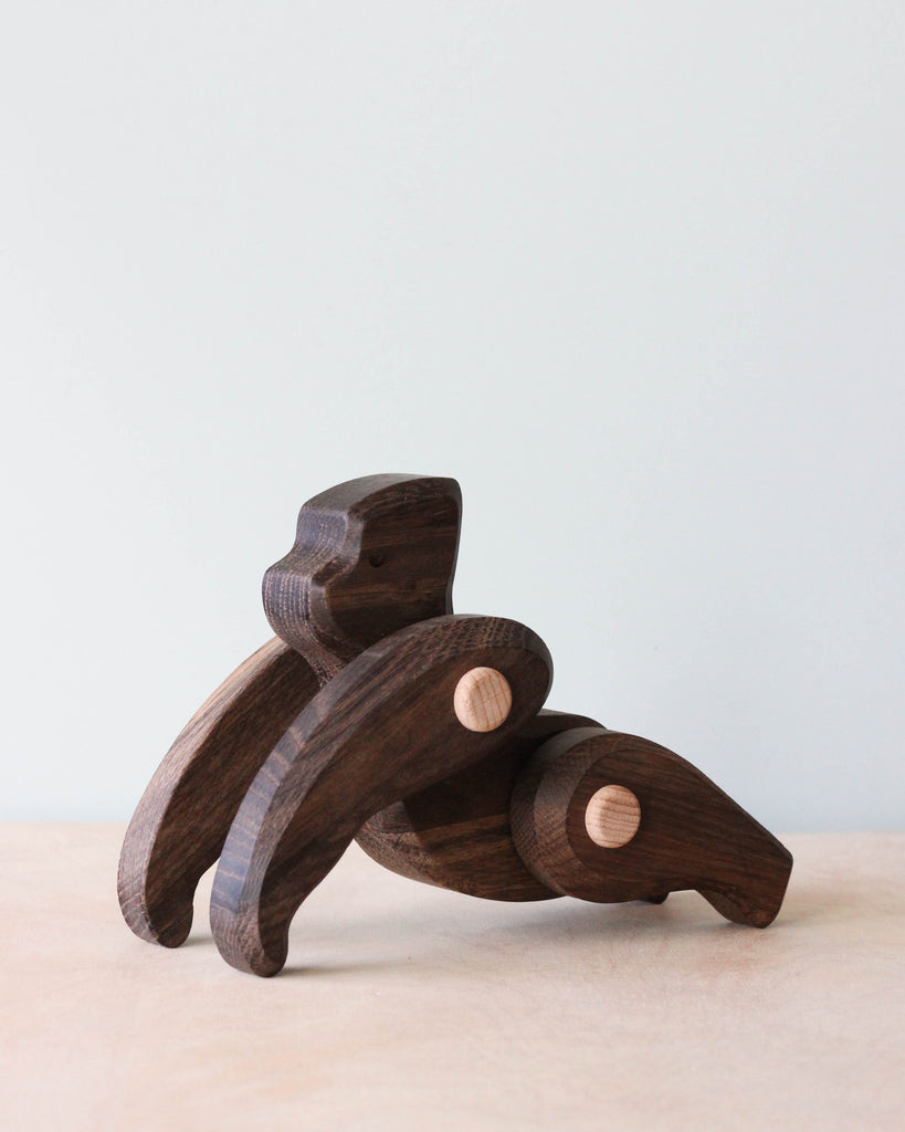 A handmade wooden gorilla with movable joints, crafted from dark wood and adorned with circular light wooden connectors. Designed for safe play using non-toxic paints, it lies on a light surface.